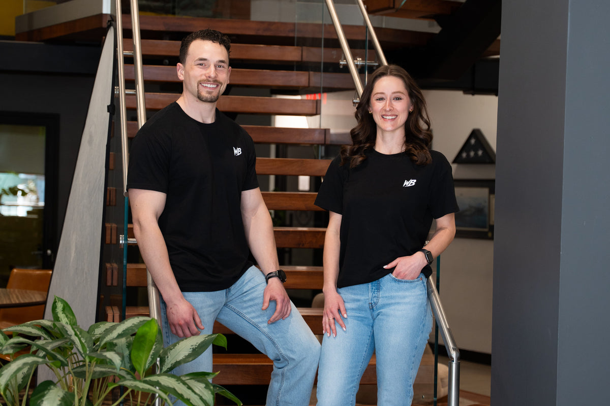 Two people wearing black t-shirts with WeBilt logo, standing in an indoor setting with stairs and plants.