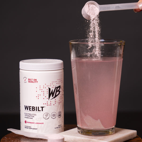 Webilt electrolyte powder being added to a glass of water on a wooden stand with a dark background.