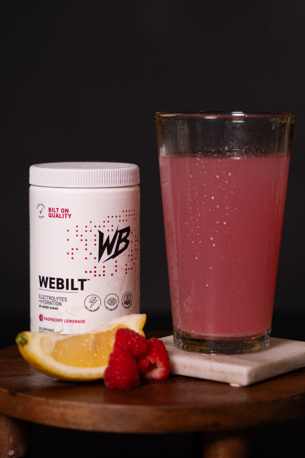 WeBilt electrolyte container with a glass of  it mixed in water and lemon and raspberry slices on a dark background