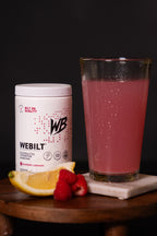 WeBilt electrolyte container with a glass of  it mixed in water and lemon and raspberry slices on a dark background